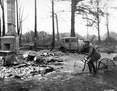 Man pretending to ride a burned-up bicycle in the ruins of Bandon, 1936